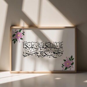 Op de afbeelding: Een ingelijste print met zwarte Arabische kalligrafie op een witte achtergrond, in een licht houten frame. Roze bloemen en groene bladeren sieren de hoeken. De print wordt gepresenteerd op een neutrale ondergrond.