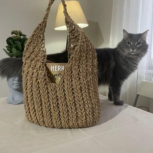 May include: A brown crocheted tote bag with a book inside. The bag has a wide, open top and a single strap. The book has the word "HERK" printed on the cover.