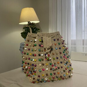 Op de afbeelding: Een beige gehaakte tote bag met kleurrijke houten kralen. De tas heeft een trekkoord sluiting en is versierd met kralen in verschillende kleuren, waaronder rood, groen, blauw, geel en wit.