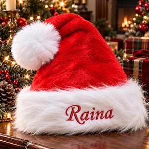 Puede incluir: Un gorro de Papá Noel de felpa roja brillante con un borde grueso de piel sintética blanca y un gran pompón blanco en la punta. La banda de piel blanca está bordada con el nombre Raina en letras rojas de estilo cursivo.