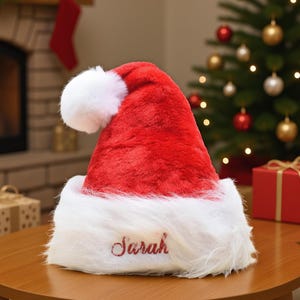 Könnte beinhalten: Ein roter Weihnachtsmannhut aus Plüsch mit weißem, flauschigem Besatz und Bommel. Der Name "Sarah" ist in roter Schrift auf den weißen Besatz gestickt. Der Hut liegt auf einem Holztisch, im Hintergrund ein Weihnachtsbaum und Geschenke.