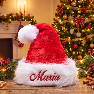 May include: A plush red Santa hat with a white faux fur trim and a white pom-pom on top. The name Maria is embroidered in red cursive script on the front white trim. The hat is made of a soft, fuzzy material and features a classic pointed shape.
