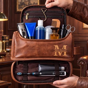 Könnte beinhalten: Eine braune Kulturbeutel aus Leder mit Tragegriff und Aufhängehaken. Auf der Vorderseite ist der Name Michael in einer Serifenschrift eingraviert. Die Tasche ist geöffnet und zeigt verschiedene Pflegeartikel wie Rasierer und Flaschen.