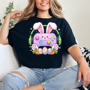 Könnte beinhalten: Schwarzes T-Shirt mit einem Cartoon-Design eines lila Gamecontrollers mit Hasenohren und Kopfhörern. Bunte Ostereier und Karotten befinden sich am unteren Rand des Controllers. Das Design ist im Cartoon-Stil gehalten.