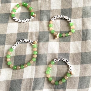Peut inclure: Quatre bracelets de perles avec le nom "Jeremiah" épelé avec des perles de lettres blanches. Les bracelets combinent des perles vertes et pêche, et de petits coquillages. Ils sont disposés sur un tissu à carreaux.