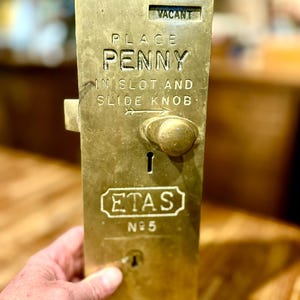 Puede incluir: Un mecanismo de cerradura de puerta rectangular de latón operado por monedas con el texto VACANT, PLACE PENNY IN SLOT AND SLIDE KNOB, ETAS y N°5. El dispositivo incluye una ranura para monedas, una perilla deslizante y una cerradura.