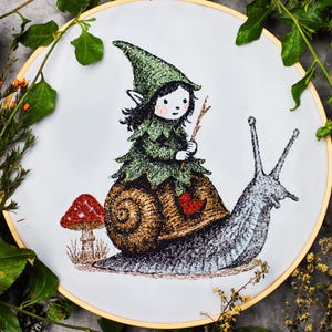Può includere: Ricamo in un cerchio di legno. Raffigura un piccolo elfo in un abito verde e frondoso che cavalca una lumaca, tenendo un ramoscello. Un fungo rosso è accanto alla lumaca. Il disegno è su sfondo bianco.