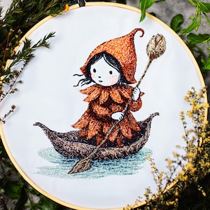 Può includere: Opera d'arte ricamata in un telaio di legno. Il disegno presenta una figura stravagante in una barca a foglia, che indossa un abito e un cappello marroni a forma di petalo, con un lungo remo a forma di cucchiaio. Sfondo bianco, con fogliame verde e giallo intorno al telaio.