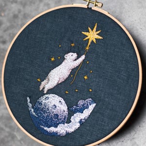 Peut inclure: Un tambour à broder circulaire en bois contenant un tissu bleu foncé avec un motif cousu à la main. La broderie représente un ours polaire blanc atteignant une grande étoile jaune, entouré de petites étoiles jaunes, d'une lune et de nuages blancs.