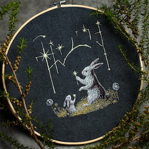May include: Embroidered artwork featuring two rabbits in a field with stars and celestial elements. The rabbits are in shades of gray and white, set against a dark gray background. The embroidery is framed in a wooden hoop.