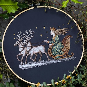 Könnte beinhalten: Gesticktes Kunstwerk auf schwarzem Hintergrund, das eine Frau in einem grünen und goldenen Kleid zeigt, die einen Schlitten fährt, der von zwei Rentieren gezogen wird. Das Design umfasst goldene Sterne und ist in einem Holzreifen gerahmt.