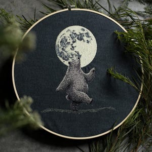 May include: Embroidered hoop art featuring a black bear dancing under a full moon. The moon is embroidered in white thread on a dark gray fabric.