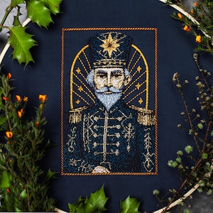May include: An embroidered portrait of a distinguished figure in a dark blue uniform with gold accents, a white beard, and a star-shaped emblem on his hat. The artwork is framed in a wooden hoop, with decorative greenery surrounding it.