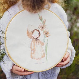 Könnte beinhalten: Ein runder Stickrahmen aus Holz mit weißem Stoff, der ein handgesticktes Motiv eines kleinen Wesens mit Kapuzenumhang zeigt, das eine hohe, hellbraune Gladiolenblüte mit grünen Stielen und Blättern auf einem schlichten Untergrund hält.