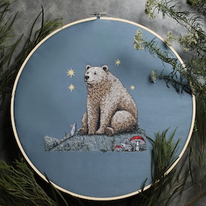 May include: Embroidered hoop art featuring a brown bear sitting on a rock with two small animals and red mushrooms. The background is a blue sky with gold stars.