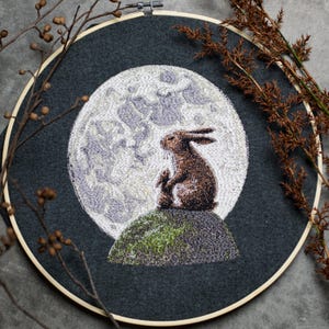 Könnte beinhalten: Stickerei in einem runden Rahmen mit einem braunen Kaninchen und einem kleineren Kaninchen, die auf einem grünen Hügel sitzen, mit einem großen weißen Mond dahinter. Der Rahmen befindet sich auf einem grauen Hintergrund.