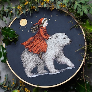 May include: An embroidered artwork featuring a girl in a red dress riding a polar bear under a night sky with a moon and stars. The embroidery is set in a wooden hoop, with a dark blue background. The artwork is surrounded by greenery.