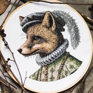 May include: Embroidered hoop art featuring a fox in Renaissance attire. The fox has brown fur, green eyes, and wears a black hat with a feather, a ruffled collar, and a green and gold patterned jacket. The artwork is set against a white background.