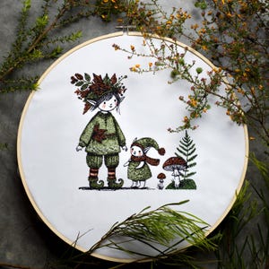 Può includere: Arte ricamata su un cerchio con due elfi, uno più grande dell'altro, con abiti verdi e stivali marroni. L'elfo più grande ha una corona di foglie sulla testa. L'elfo più piccolo indossa una sciarpa rossa. Entrambi gli elfi sono in piedi accanto a un fungo e una felce.