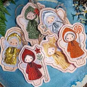 May include: A selection of handmade angel decorations with embroidered details. Each decoration depicts a child-like figure in a hooded robe, with wings and a sweet expression. The robes are in various colours, including gold, red, and green.