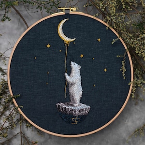 May include: A circular wooden embroidery hoop containing a dark blue fabric background. The embroidery depicts a white polar bear standing on a small icy patch, holding a thin yellow thread attached to a crescent moon with yellow stars.