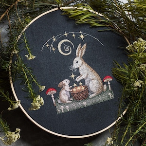 May include: Embroidered hoop art featuring two white rabbits, one larger than the other, sitting in front of a small drum. The rabbits are surrounded by small red mushrooms and gold stars. The background is a dark blue fabric.