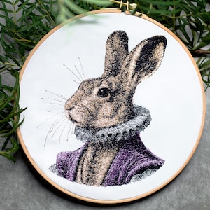 Puede incluir: Arte de bordado en aro con un conejo marrón que lleva un cuello de color morado y blanco. Los bigotes del conejo están detallados con hilo blanco.