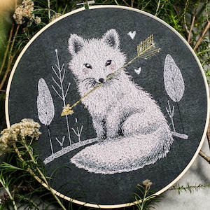 May include: Embroidered hoop art featuring a white arctic fox with a gold arrow through its mouth, surrounded by stylised trees on a dark grey background.