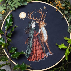 May include: Embroidered artwork on black fabric, featuring a woman in a red dress and green cloak with a deer. A full moon and stars are in the background. The artwork is framed in a wooden hoop and surrounded by greenery.