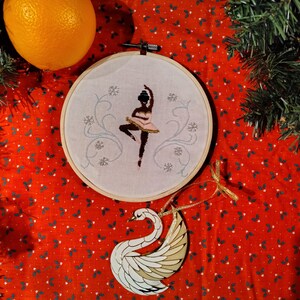 May include: A white swan ornament with a gold design, a red fabric background with white holly leaves, and a white embroidery hoop with a black ballerina embroidered on it.