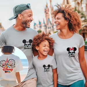 Pu&ograve; includere: Una famiglia indossa magliette grigie abbinate con disegni di Topolino e Minnie. Le magliette presentano nomi personalizzati e un design Disney 2023 sul retro di una maglietta. La famiglia sorride davanti a un parco a tema.