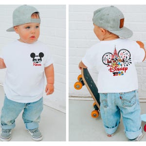 Pu&ograve; includere: Maglietta bianca con grafica di Topolino con occhiali da sole e il nome "Noah" in rosso. Il retro della maglietta presenta un design Disney 2023 con orecchie di Topolino, un castello e personaggi. Il bambino indossa un berretto da baseball grigio e jeans blu.