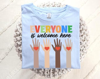 May include: Light blue t-shirt with the text "EVERYONE is welcome here" in rainbow colors. The text is above four stylized hands with hearts in the palms.