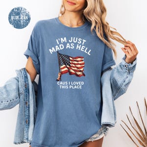 May include: Blue t-shirt with the text "I'M JUST MAD AS HELL CAUS I LOVED THIS PLACE" and an American flag graphic. The shirt is a light blue color and has rolled-up sleeves. A "BLUE JEAN" logo is in the upper left corner.