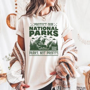 May include: White t-shirt with a green graphic design that says "Protect Our National Parks" with a mountain scene and the text "Defund Doge Parks, Not Profits".