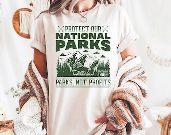 May include: White t-shirt with a green graphic design that says "Protect Our National Parks" with a mountain scene and the text "Defund Doge Parks, Not Profits".