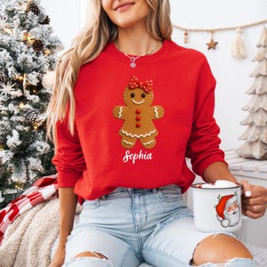 Op de afbeelding: Een rode sweatshirt met een peperkoeken vrouw ontwerp, een rode strik en drie rode knopen. De naam "Sophia" is in witte letters onder de peperkoeken figuur gedrukt. De persoon houdt een witte mok vast met een illustratie van de kerstman.