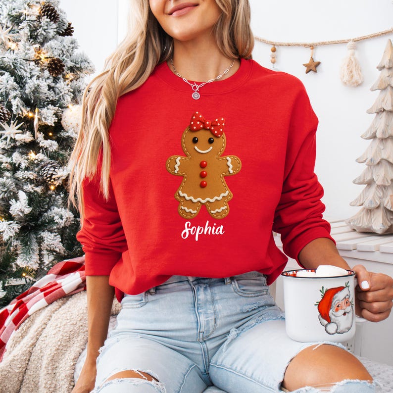 Op de afbeelding: Een rode sweatshirt met een peperkoeken vrouw ontwerp, een rode strik en drie rode knopen. De naam "Sophia" is in witte letters onder de peperkoeken figuur gedrukt. De persoon houdt een witte mok vast met een illustratie van de kerstman.
