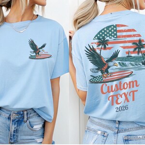 May include: Light blue t-shirt featuring an eagle on a surfboard with an American flag design. The front has a small graphic of the eagle and surfboard. The back has a larger design with the text "Custom TEXT 2026".