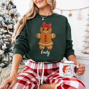 Op de afbeelding: Een donkergroene sweatshirt met een peperkoekenmannetje, een rode strik en de naam "Emily". De persoon draagt een rood-witte geruite pyjamabroek en houdt een mok met een kerstman-afbeelding vast.