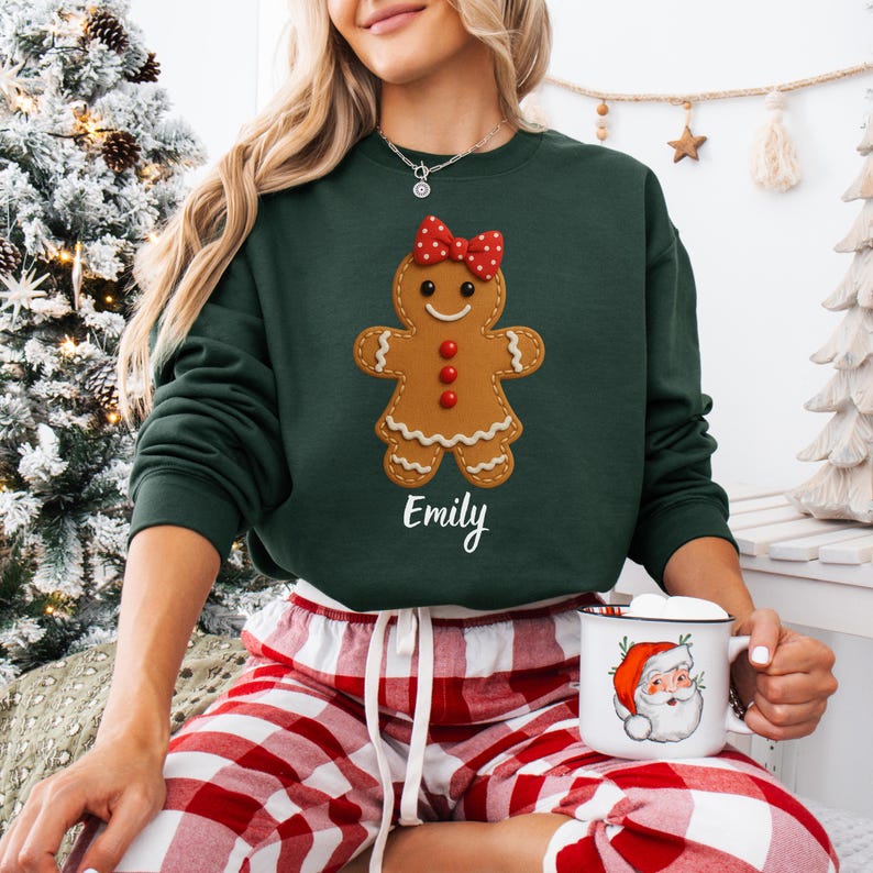 Op de afbeelding: Een donkergroene sweatshirt met een peperkoekenmannetje, een rode strik en de naam "Emily". De persoon draagt een rood-witte geruite pyjamabroek en houdt een mok met een kerstman-afbeelding vast.