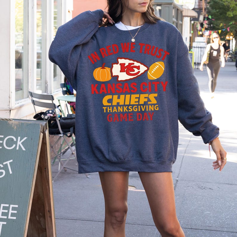 Puede incluir: Una sudadera azul marino con el texto "IN RED WE TRUST" y "KANSAS CITY CHIEFS THANKSGIVING GAME DAY" en rojo y amarillo. El dise&ntilde;o incluye una calabaza, un bal&oacute;n de f&uacute;tbol americano y el logotipo de los Chiefs.