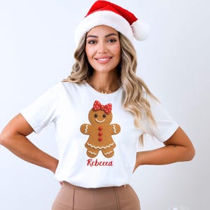 Op de afbeelding: Wit T-shirt met een peperkoekenmannetje, een rode strik en knopen. De naam "Rebecca" staat in het rood onder het peperkoekenmannetje. De persoon op de foto draagt een kerstmuts.
