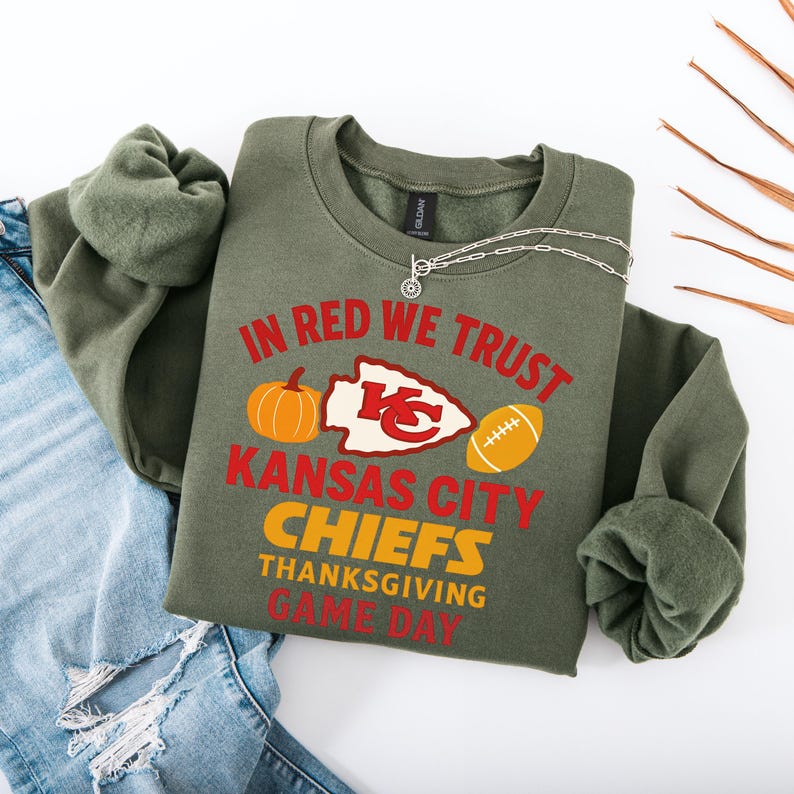 Puede incluir: Una sudadera verde oliva con el texto "IN RED WE TRUST" y "KANSAS CITY CHIEFS THANKSGIVING GAME DAY" en rojo y amarillo. El dise&ntilde;o incluye una calabaza, un bal&oacute;n de f&uacute;tbol y el logotipo de los Chiefs. Un collar plateado est&aacute; drapeado sobre la sudadera.