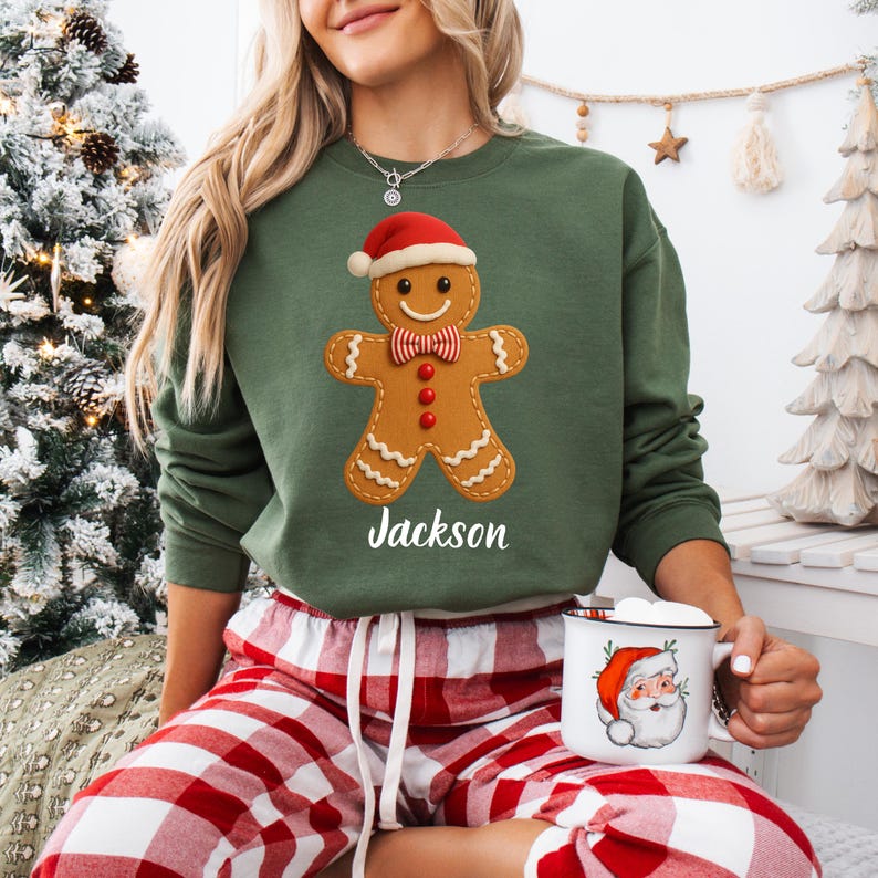 Op de afbeelding: Groen sweatshirt met een peperkoekmannetje met een kerstmuts en een vlinderdas. De naam "Jackson" staat eronder gedrukt. De persoon draagt een rood-witte geruite pyjamabroek en houdt een mok met een kerstmanontwerp vast.