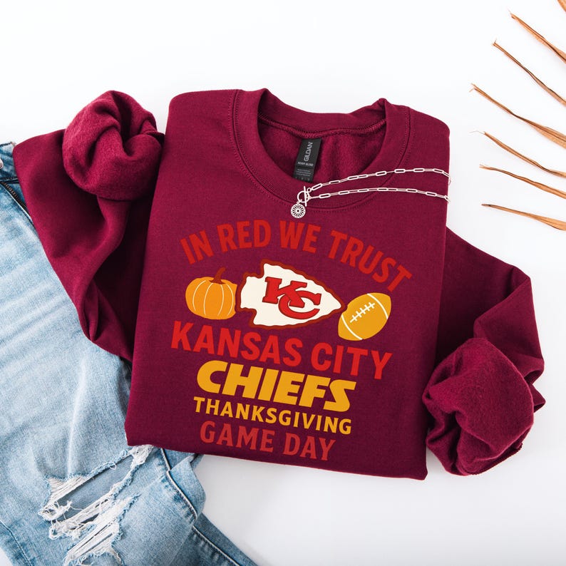 Puede incluir: Sudadera burdeos con la frase "IN RED WE TRUST" y "KANSAS CITY CHIEFS THANKSGIVING GAME DAY". El dise&ntilde;o incluye una calabaza, un bal&oacute;n de f&uacute;tbol y el logotipo de los Chiefs. Un collar plateado est&aacute; sobre la sudadera.