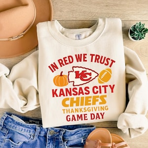 May include: Cream-colored sweatshirt with the text "IN RED WE TRUST" and "KANSAS CITY CHIEFS THANKSGIVING GAME DAY" in red and orange. The design includes a pumpkin, a football, and the Chiefs logo.