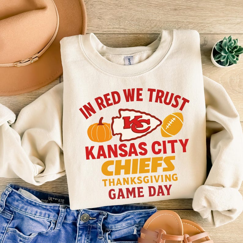 Puede incluir: Sudadera color crema con el texto "IN RED WE TRUST" y "KANSAS CITY CHIEFS THANKSGIVING GAME DAY" en rojo y naranja. El dise&ntilde;o incluye una calabaza, un bal&oacute;n de f&uacute;tbol americano y el logo de los Chiefs.