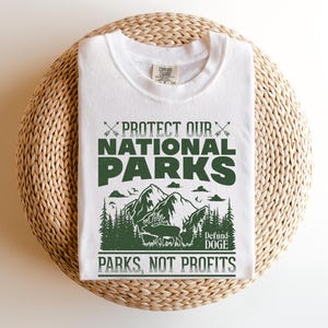 May include: White t-shirt with a green graphic design that says "Protect Our National Parks" with a mountain scene and the text "Defund Doge Parks, Not Profits".
