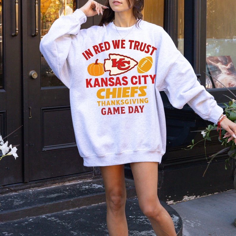 Puede incluir: Una sudadera blanca con el texto "IN RED WE TRUST" sobre un logotipo de los Kansas City Chiefs, una calabaza y un bal&oacute;n de f&uacute;tbol. Debajo, se lee "KANSAS CITY CHIEFS THANKSGIVING GAME DAY".